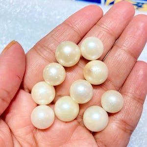 May include: A collection of lustrous, creamy-white pearls, each with a smooth, spherical shape. The pearls are held in a hand, showcasing their natural iridescence and subtle variations in color. These pearls are ideal for jewelry making.
