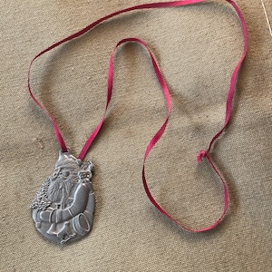Könnte beinhalten: Silberfarbener Weihnachtsschmuck mit Santa Claus, der einen Baum und eine Trommel hält, mit einem roten Band zum Aufhängen. Der Schmuck ist mit geätzten Designs versehen und eignet sich für die Weihnachtsdekoration.