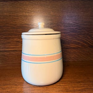 Pode incluir: Um pote de cerâmica creme com tampa, com uma faixa rosa entre duas faixas azuis. O pote tem formato arredondado e um pequeno botão na tampa. O pote é provavelmente para armazenamento na cozinha.