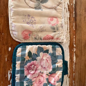 Puede incluir: Dos agarraderas con estampado floral. Una tiene un fondo crema con rosas rosas y un diseño de ramas. La otra tiene un patrón de cuadros azules y blancos con rosas rosas y un borde verde azulado. Las agarraderas son cuadradas con esquinas redondeadas.