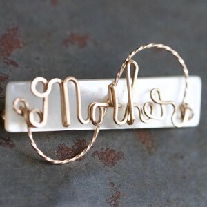 Puede incluir: Una base rectangular blanca con la palabra "Madre" escrita en alambre dorado. El alambre está retorcido y forma un corazón al final de la palabra.