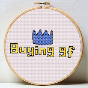 Può includere: Ricamo a punto croce in un telaio di legno. Il disegno presenta una corona blu sopra le parole "Buying gf" in lettere gialle. Lo sfondo è un tessuto rosa chiaro.