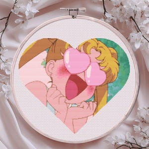 May include: A cross-stitch embroidery hoop featuring a cartoon character with heart-shaped eyes and a surprised expression. The design is set within a heart shape, with a pink and green color scheme. The hoop is set against a soft, white background with floral accents.