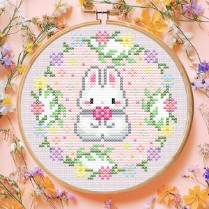Puede incluir: Un bastidor de bordado de punto de cruz con un conejito blanco sosteniendo un corazón rosa, rodeado por una corona floral. El diseño está sobre un fondo rosa claro, con un aro de madera. La imagen está enmarcada por flores coloridas.