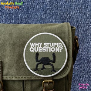 May include: An olive green messenger bag with a round patch that reads "WHY STUPID QUESTION?" in white lettering. The patch features a black creature design. The bag has brown leather straps and buckles.