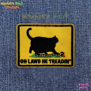 May include: Embroidered patch featuring a black cat stepping on a snake, with the text "OH LAWD HE TREADIN" on a yellow background. The patch has a black border and is set against a denim background.