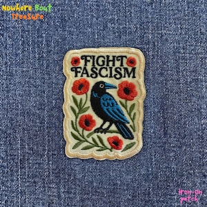 May include: Embroidered patch with the words "FIGHT FASCISM" above a blue bird illustration surrounded by red poppies and green leaves. The patch has a cream-colored background and a stitched border. The patch is iron-on.