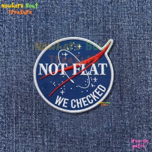 May include: A circular iron-on patch with a blue background and white border. The patch features the text "NOT FLAT WE CHECKED" in white, with a red and white graphic. The patch is designed to be ironed onto fabric.