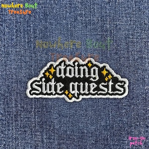 May include: An iron-on patch with the words "doing side quests" in white script on a black background. The patch is outlined in white with yellow star accents. The patch is on a blue denim background.