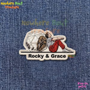 May include: An iron-on patch featuring a person in red pants and a gray shirt, leaning against a geometric structure with a brown figure inside. The text "Rocky & Grace" is below the image.