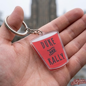 May include: A clear acrylic keychain with a red cup design. The keychain features the words "DUKE AND RALLY" in black lettering. The keychain is attached to a silver metal keyring and chain.
