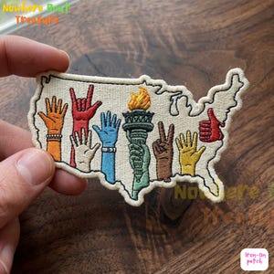 May include: Embroidered patch in the shape of the United States, featuring various hand gestures and a torch. The patch includes the text "Nowhere But Treasure" and "Iron-on patch". The hands are in various colors, including orange, red, blue, brown, and yellow.