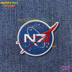 May include: Embroidered patch featuring a blue circle with a white border, the letters "N" and "Z" in white, and a red swoosh. The patch has a NASA-inspired design with stars and a white outline. The text "iron-on patch" is visible.