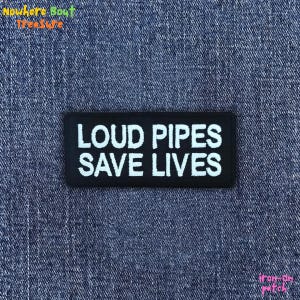 May include: A black rectangular iron-on patch with the white text "LOUD PIPES SAVE LIVES". The patch is displayed on a blue denim background. The patch is a motorcycle or biker-themed accessory.