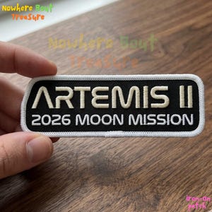 May include: A black and white rectangular patch with the text "ARTEMIS II 2026 MOON MISSION". The patch has a white border and is held by a hand. The patch is an iron-on patch.