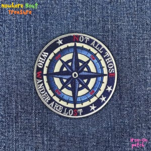 May include: Round embroidered patch with a compass design in shades of blue, white, and red. The text "NOT ALL THOSE WHO WANDER ARE LOST" is displayed around the compass. The patch is on a denim background.