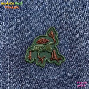 May include: An iron-on patch featuring a cartoonish creature with a brown head, green body, and orange legs and arms, holding a trident. The patch is outlined in green and has the text "Nowhere Bout Treasure" at the top.