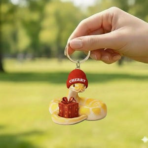 May include: A yellow and white snake-shaped keychain wearing a red beanie with the word "CHERRY" and holding a red gift box with a bow. The keychain is attached to a silver ring.