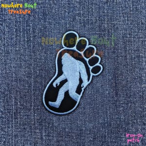 May include: An iron-on patch featuring a light blue Bigfoot silhouette inside a black footprint outline. The patch has a light blue border and the text "iron-on patch" is visible in the bottom right corner.