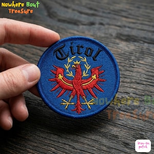 May include: Round blue embroidered patch with the word "Tirol" arched across the top. A red eagle with yellow accents is centered below the text. The patch is an iron-on style.