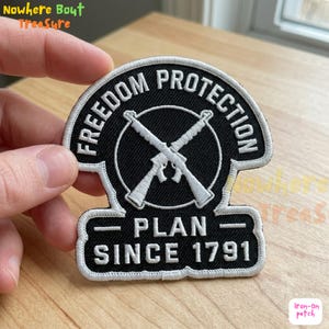 Puede incluir: Parche bordado en blanco y negro con las palabras "FREEDOM PROTECTION" y "PLAN SINCE 1791". El parche presenta un diseño circular con rifles cruzados en el centro. El parche es para planchar.