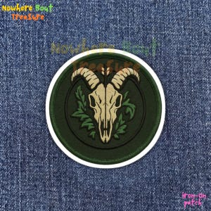 May include: Round iron-on patch featuring a goat skull with large horns, surrounded by green leaves, set against a dark green background. The patch has a white border and the text "iron-on patch" is visible at the bottom.