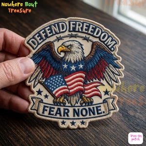 May include: Embroidered patch featuring an American bald eagle with outstretched wings, the US flag, and the text "DEFEND FREEDOM" and "FEAR NONE." The patch has a beige border and is in red, white, blue, and gold.