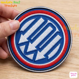 May include: Round iron-on patch with a blue center featuring a white design of arrows and lines. The patch is bordered by red, white, and black rings. The text "Nowhere Bout Treasure" is visible at the top left.
