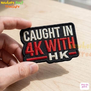 May include: Black rectangular iron-on patch with white and red embroidered text. The text reads "CAUGHT IN 4K WITH HK-". The patch has a black border and a red horizontal line.