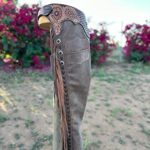 Peut inclure: Chaps en daim marron avec des détails floraux et géométriques en cuir travaillé à la taille. Comprend des boutons argentés décoratifs, des franges et une poche. Conçus pour l'équitation.