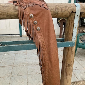 May include: Brown leather chaps with fringe and decorative silver-toned buckles. The top section features intricate floral tooling. The chaps are hanging on a wooden rail, showcasing their design and craftsmanship. A western-style garment.