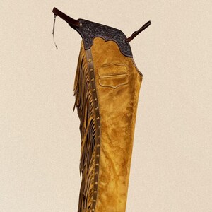 May include: Mustard yellow suede chaps with brown leather detailing and fringe. The chaps have an embossed leather yoke and a pocket. Western wear.