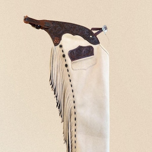 Puede incluir: Chaps de cuero color hueso con un canesú de cuero marrón oscuro que presenta un grabado floral detallado y el número 80 en relieve. La pierna incluye largos flecos a juego, conchos metálicos decorativos a lo largo de la costura y un parche de bolsillo de cuero marrón oscuro.