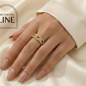 Peut inclure: Une bague dorée avec un motif de maillons de chaîne, ornée de petites pierres scintillantes. La bague est portée sur une main avec une manucure aux tons neutres. L'image comprend le texte "THE CURATED LINE" en noir.