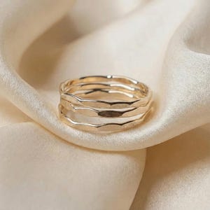May include: A stack of four gold-colored rings with a textured, organic design. The rings are displayed on a cream-colored, silky fabric, creating a soft and elegant aesthetic. The rings appear to be made of metal.