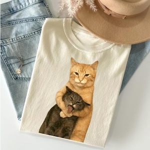 May include: Cream-colored t-shirt featuring a graphic of two cats embracing. One cat is orange, the other is gray. The shirt is paired with light blue jeans, a necklace, and a tan hat.