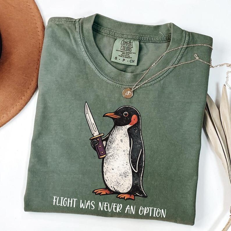 Pode incluir: Uma t-shirt de gola redonda verde-oliva com um gr&aacute;fico de um pinguim segurando uma faca. O texto abaixo do pinguim diz "FLIGHT WAS NEVER AN OPTION". A t-shirt tem uma etiqueta da marca Comfort Colors dentro da gola, indicando o tamanho S.