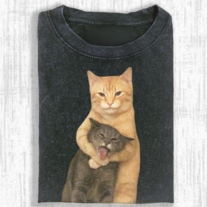 May include: Black t-shirt featuring a graphic of two cats. One orange tabby cat is hugging a gray cat, which has its mouth open. The t-shirt is folded and has a crew neck.