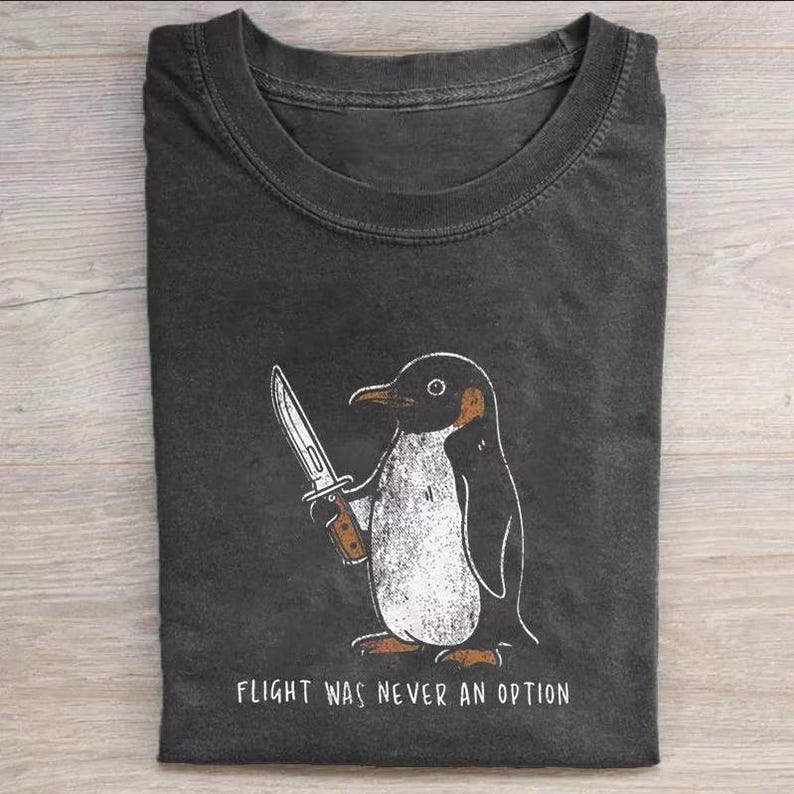 Pode incluir: Uma t-shirt de gola redonda cinzento-carv&atilde;o com uma ilustra&ccedil;&atilde;o gr&aacute;fica de um pinguim a segurar uma faca. Abaixo do pinguim, o texto diz "FLIGHT WAS NEVER AN OPTION" em letras mai&uacute;sculas brancas. O design tem um aspeto texturado e desgastado.