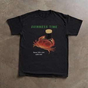May include: Black t-shirt with a graphic of a crab holding a pint of Guinness. The text "GUINNESS TIME" is above the crab, and "Have this one with me!" is below. The crab is brown and the beer has a tan head.