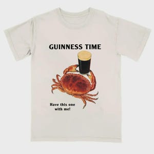 May include: Cream-coloured t-shirt with a crab illustration holding a pint of stout. The text "GUINNESS TIME" is above the crab, and "Have this one with me!" is below. The crab is brown and red, and the beer has a creamy head.