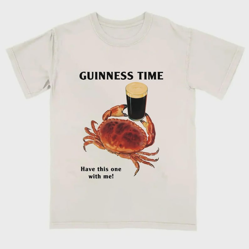 May include: Cream-coloured t-shirt with a crab illustration holding a pint of stout. The text "GUINNESS TIME" is above the crab, and "Have this one with me!" is below. The crab is brown and red, and the beer has a creamy head.