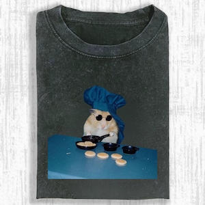 May include: Dark gray t-shirt featuring a humorous image of a hamster wearing a chef's hat and sunglasses, cooking pancakes. The hamster is positioned behind a blue table with small black pans and pancakes.
