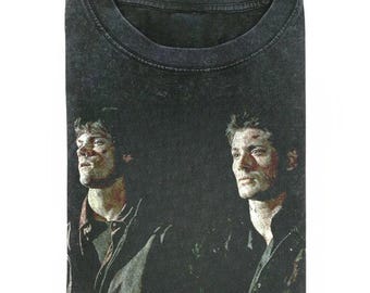 May include: A black acid-wash t-shirt featuring a graphic print of two men with short hair, both wearing dark jackets. The print shows the individuals from the chest up, with one man looking to the left and the other looking forward.