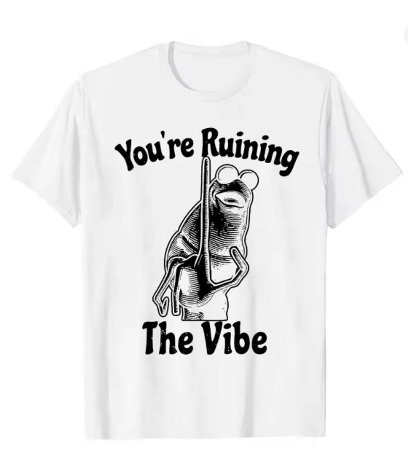 Puede incluir: Camiseta blanca con una ilustraci&oacute;n en blanco y negro de una rana con gafas sosteniendo un poste. El texto "You're Ruining The Vibe" est&aacute; arriba y abajo del gr&aacute;fico de la rana. La camiseta tiene mangas cortas y cuello redondo.