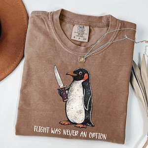 Pode incluir: Uma t-shirt castanha de manga curta com um gr&aacute;fico de um pinguim a segurar uma faca. O texto abaixo do pinguim diz "FLIGHT WAS NEVER AN OPTION". A t-shirt tem uma etiqueta da marca Comfort Colors no pesco&ccedil;o, indicando o tamanho S.