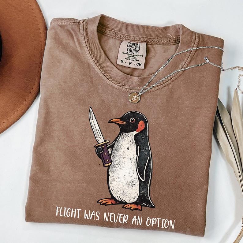 Pode incluir: Uma t-shirt castanha de manga curta com um gr&aacute;fico de um pinguim a segurar uma faca. O texto abaixo do pinguim diz "FLIGHT WAS NEVER AN OPTION". A t-shirt tem uma etiqueta da marca Comfort Colors no pesco&ccedil;o, indicando o tamanho S.