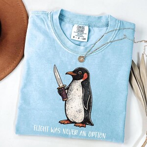 Pode incluir: Uma t-shirt azul claro com um gr&aacute;fico de um pinguim a segurar uma faca e o texto "FLIGHT WAS NEVER AN OPTION" impresso por baixo. A pe&ccedil;a tem uma etiqueta da marca Comfort Colors no decote interior e um design de gola redonda.
