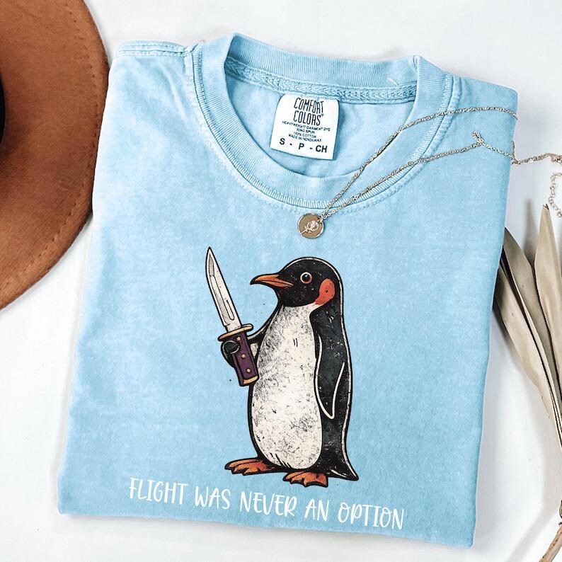Pode incluir: Uma t-shirt azul claro com um gr&aacute;fico de um pinguim a segurar uma faca e o texto "FLIGHT WAS NEVER AN OPTION" impresso por baixo. A pe&ccedil;a tem uma etiqueta da marca Comfort Colors no decote interior e um design de gola redonda.