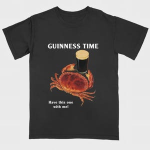 May include: Black t-shirt with a crab graphic holding a pint of stout. The text "GUINNESS TIME" is above the crab, and "Have this one with me!" is below. The crab is red-brown, and the beer has a tan head.